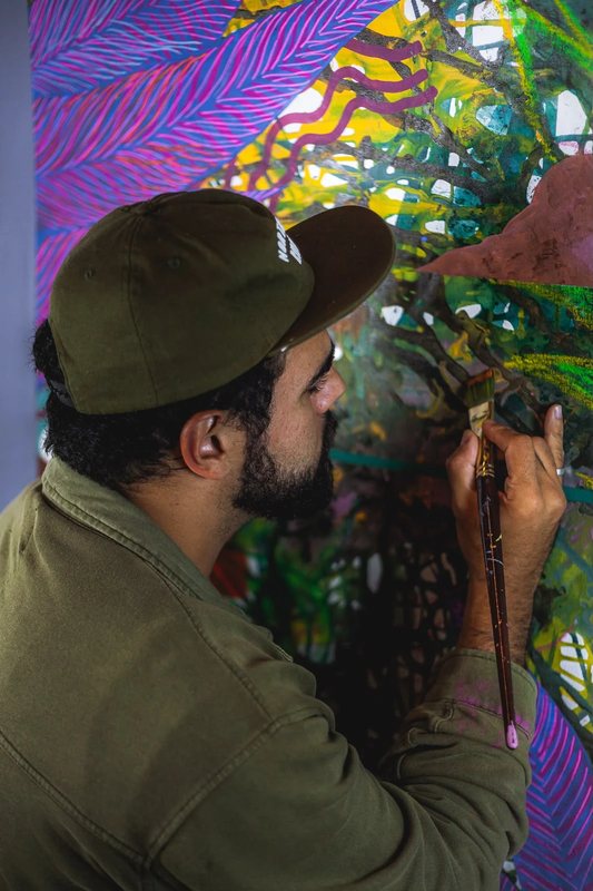João Ferreira de boné, de perfil, pintando com um pincel largo uma tela de folhas coloridas.
